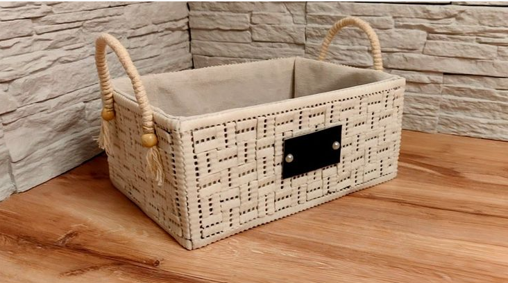 A woven cream-colored rectangular storage basket featuring braided rope handles with decorative beads, a fabric lining inside, and a dark rectangular plaque on the front, exemplifying practical home decor ideas without buying anything by organizing and repurposing storage containers.