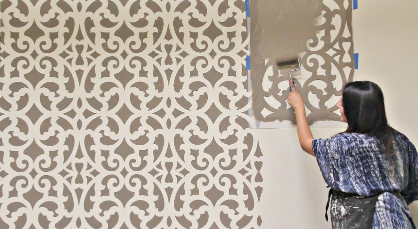 A woman using a paint roller and a large decorative wall stencil to create an intricate pattern, representing great crafts for people who arent artistic.