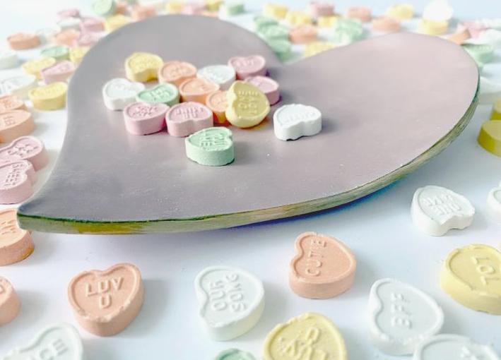 A light purple heart-shaped ceramic dish holding small pastel candy hearts, showcasing simple crafts for people who arent artistic.