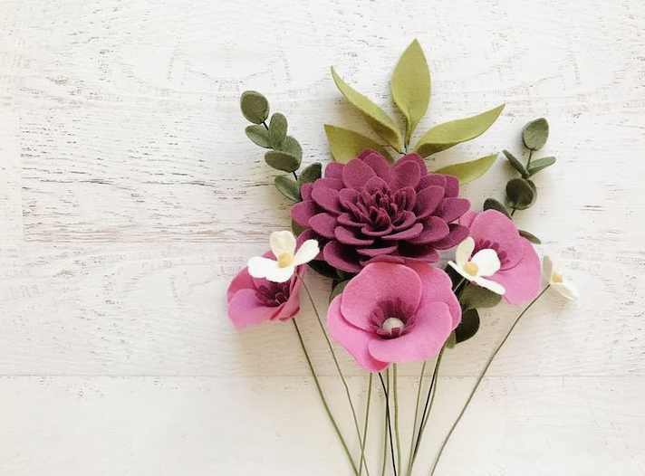 A bouquet of handcrafted felt flowers in shades of deep plum and pink, accompanied by green leaves on stems against a white wooden background, making it a wonderful example of home decor ideas without buying anything.