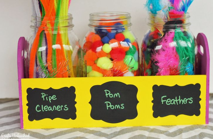 Three glass mason jars filled with brightly colored pipe cleaners, colorful pom poms, and vibrant craft feathers, organized inside a yellow wooden caddy with chalkboard labels, demonstrating practical home decor ideas without buying anything.