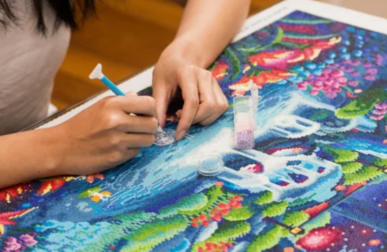 A person using a specialized tool to place small resin rhinestones onto a vibrant, colorful diamond painting canvas, representing creative home decor ideas without buying anything.