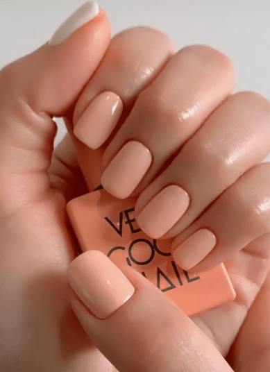 25 Nail Colors That Make Hands Look Younger A close-up of short, squared peach-toned nails holding a matching polish bottle, demonstrating nail colors that make hands look younger.
