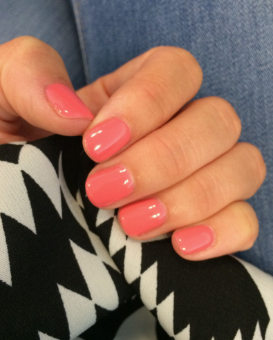 25 Nail Colors That Make Hands Look Younger A close-up of a hand resting on a black-and-white patterned fabric, featuring bright coral-pink square nails that showcase vibrant nail colors that make hands look younger.