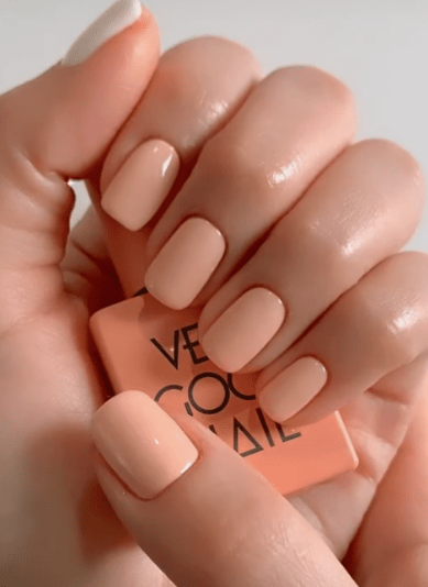 25 Nail Colors That Make Hands Look Younger A close-up of short, squared peach-toned nails holding a matching polish bottle, demonstrating nail colors that make hands look younger.