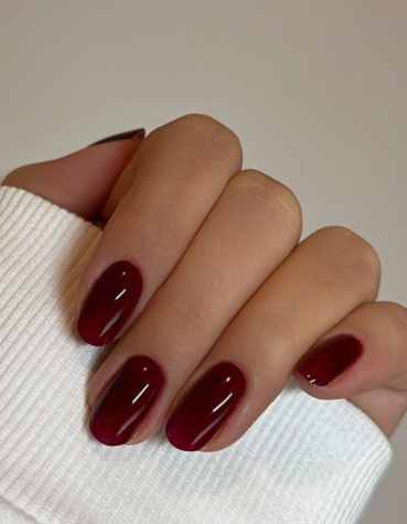25 Nail Colors That Make Hands Look Younger A hand wearing a white sleeve showcasing rich glossy burgundy oval nails as one of the chic nail colors that make hands look younger.
