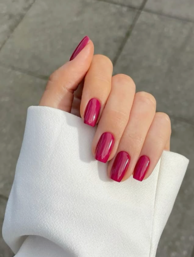 25 Nail Colors That Make Hands Look Younger A hand wearing a white sleeve showcasing glossy magenta-berry square nails, highlighting vibrant nail colors that make hands look younger.