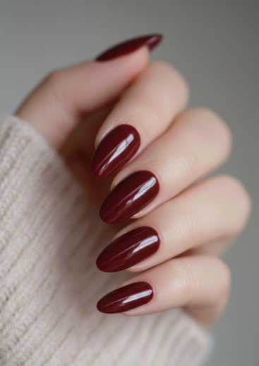 25 Nail Colors That Make Hands Look Younger A hand wearing a neutral beige sweater displaying glossy dark burgundy oval nails, highlighting classic nail colors that make hands look younger.