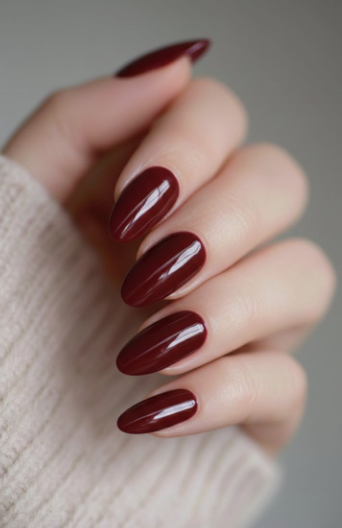 25 Nail Colors That Make Hands Look Younger A hand wearing a neutral beige sweater displaying glossy dark burgundy oval nails, highlighting classic nail colors that make hands look younger.