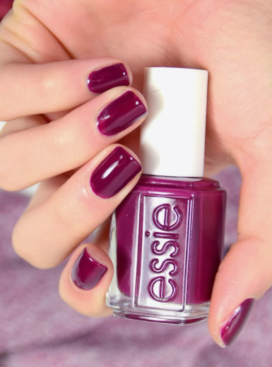 25 Nail Colors That Make Hands Look Younger A close-up of a hand holding an Essie nail polish bottle, showcasing rich plum-purple square nails as one of the best nail colors that make hands look younger