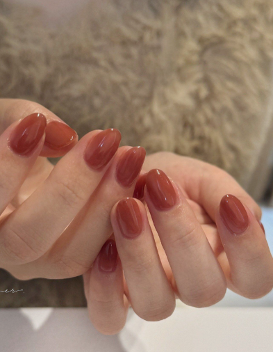 25 Nail Colors That Make Hands Look Younger A close-up of hands displaying glossy terracotta-brown oval nails, illustrating warm nail colors that make hands look younger.