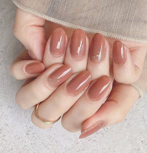 25 Nail Colors That Make Hands Look Younger A close-up of hands wearing a sheer neutral top, showcasing glossy caramel-brown oval nails with subtle sparkle accents as charming nail colors that make hands look younger.
