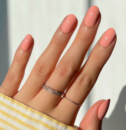 25 Nail Colors That Make Hands Look Younger A hand adorned with delicate silver rings and wearing a yellow-striped sleeve, showcasing glossy pastel-peach oval nails as vibrant nail colors that make hands look younger.
