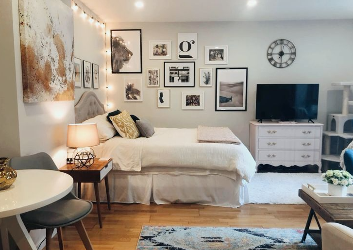 A cozy studio bedroom featuring neutral bedding, a gallery wall of framed prints, string lights, and a white dresser, showcasing smart small apartment decor ideas.