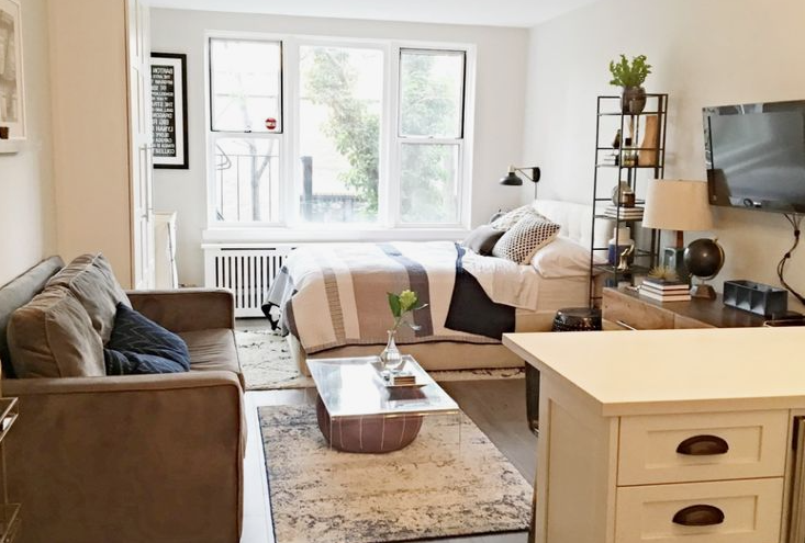 An open-concept studio apartment featuring a cozy living room with a brown sofa, a glass coffee table, and a neatly made bed near large windows, highlighting functional small apartment decor ideas.