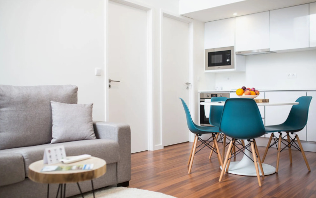 A bright, minimalist apartment living and dining area featuring a grey sofa, wooden coffee table, and a round dining table with vibrant teal chairs, showcasing sleek small apartment decor ideas.