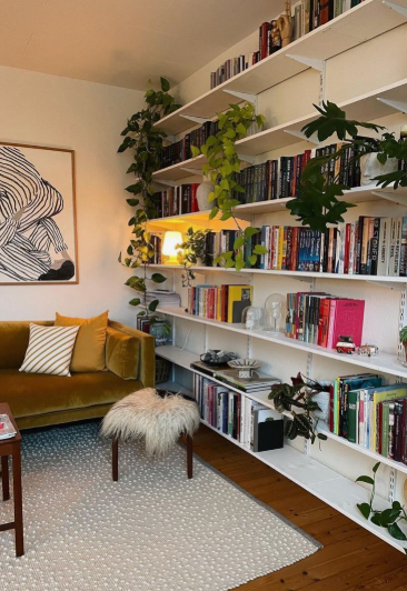 A cozy living space featuring floor-to-ceiling white wall-to-wall bookshelves overflowing with books and trailing green plants, paired with a mustard velvet sofa and a textured rug, highlighting creative small apartment decor ideas.