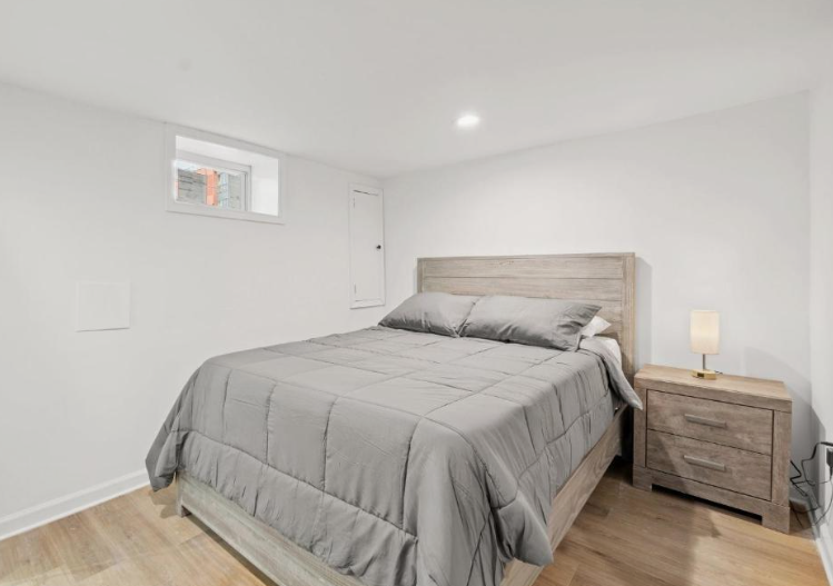 A minimalist bedroom featuring a grey wooden bed frame, matching nightstand, and a small high-set window, reflecting clean and simple small apartment decor ideas.