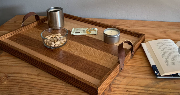 A handcrafted wooden serving tray featuring striped wood panels and leather loop handles, styled with a book, a candle, and snacks, showing a gorgeous project for DIY crafts that look expensive.