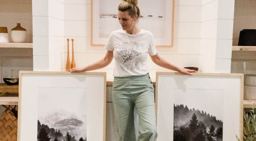 A woman holding up two large, framed black-and-white landscape prints in a modern white-paneled room, showcasing stunning DIY crafts that look expensive for upscale wall art.
