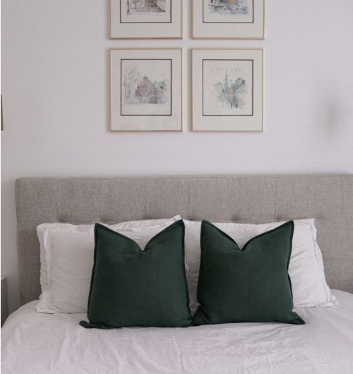 A neatly styled bed with a tufted grey headboard, crisp white pillows, and rich forest green accent cushions beneath a gallery wall of framed botanical sketches, presenting stunning DIY crafts that look expensive for bedroom decor.