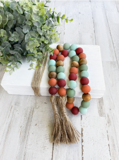 A colorful wooden bead garland in shades of red, teal, sage green, and natural wood with jute tassels, styled on a white book stack with eucalyptus, highlighting charming DIY crafts that look expensive.