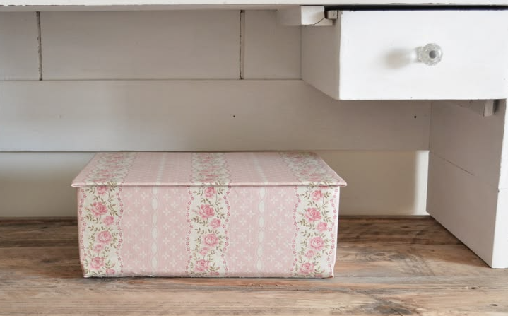 A fabric-covered storage box in a delicate pink floral vintage pattern placed neatly under a white wooden desk, showcasing charming DIY crafts that look expensive for home organization.