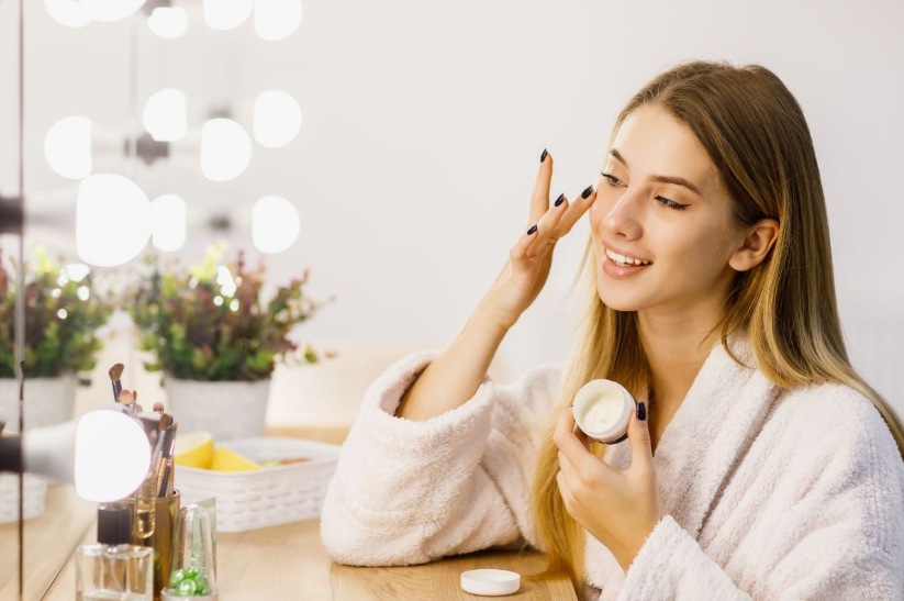 2026 Beauty Trends That Are Actually Worth Trying A smiling woman in a plush white bathrobe applying facial cream in front of a lighted vanity mirror, capturing essential self-care practices and 2026 beauty trends for skincare routines.