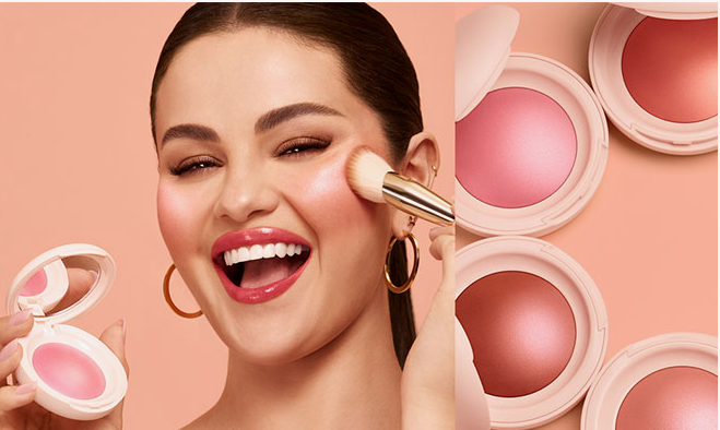 2026 Beauty Trends That Are Actually Worth Trying A promotional shot of a smiling model applying glowing blush with a makeup brush alongside open compacts of pink and peach blush, highlighting key 2026 beauty trends for luminous face makeup.