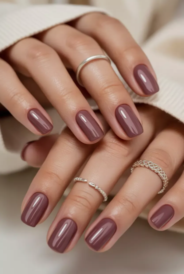30 Classy Nail Colors for Mature Hands Hands showcasing classy nail colors for mature hands, featuring short, glossy dark mauve-brown polished nails styled with delicate silver rings against a neutral fabric background.