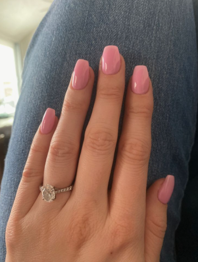 30 Classy Nail Colors for Mature Hands A hand featuring a clean, glossy bubblegum pink manicure with short coffin nails, accented with a sparkling diamond engagement ring resting against blue denim.
