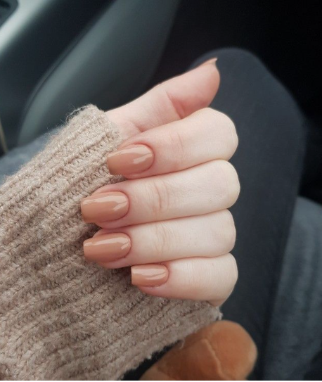 30 Classy Nail Colors for Mature Hands A hand featuring a polished, glossy nude-beige manicure with short square nails, resting against a cozy tan knitted sweater.