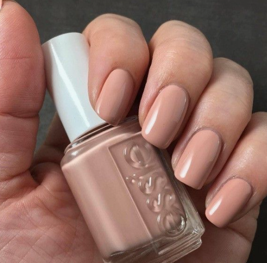 30 Classy Nail Colors for Mature Hands A hand holding an Essie nail polish bottle, showcasing classy nail colors for mature hands featuring a glossy neutral peach-beige manicure with short, soft-square nails.