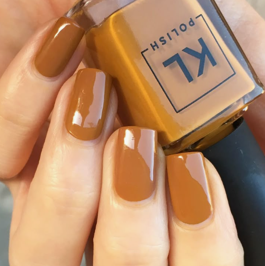 30 Classy Nail Colors for Mature Hands A hand holding a KL Polish bottle, showcasing classy nail colors for mature hands featuring a glossy caramel-tan manicure with short square nails.