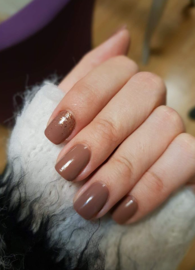 30 Classy Nail Colors for Mature Hands A hand showcasing classy nail colors for mature hands, featuring glossy brown short square nails with a sparkly gold glitter accent nail, resting against a fuzzy white sweater.
