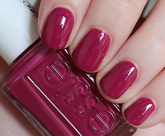 30 Classy Nail Colors for Mature Hands A hand holding an Essie nail polish bottle, showcasing classy nail colors for mature hands featuring a glossy berry-magenta manicure with short square nails.