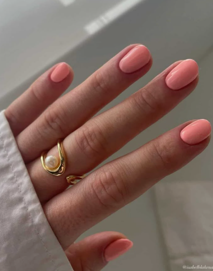 30 Classy Nail Colors for Mature Hands Hands showcasing classy nail colors for mature hands, featuring a glossy soft peach-coral manicure on short oval nails accented with a sculptural gold and pearl ring.