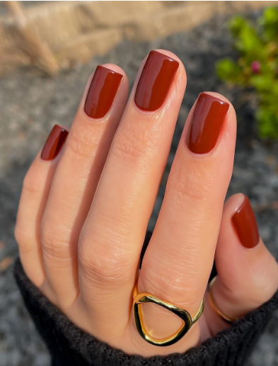 30 Classy Nail Colors for Mature Hands Hands showcasing classy nail colors for mature hands, featuring a glossy rust-red manicure on short square nails accented with a bold gold statement ring.