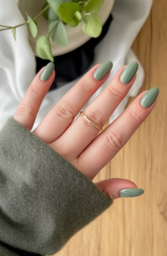 30 Classy Nail Colors for Mature Hands Hands showcasing classy nail colors for mature hands, featuring a glossy sage green oval manicure paired with a minimalist gold band and a matching green sweater sleeve.
