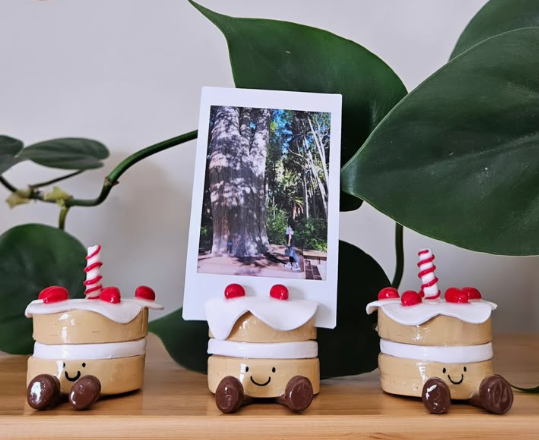 Three cute air dry clay ideas shaped like smiling miniature cakes with red cherries and striped candles, with the center cake holding a Polaroid photo in front of a green plant.