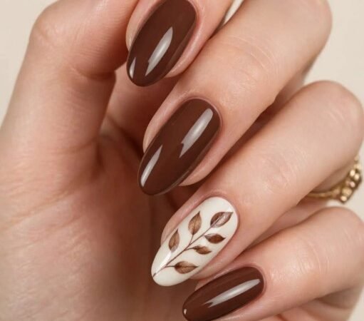 Fall Nail Inspo 2026: 20 Cozy & Trendy Designs Fall nail inspo 2026 featuring glossy chocolate brown almond nails with a cream accent nail painted with brown autumn leaves.