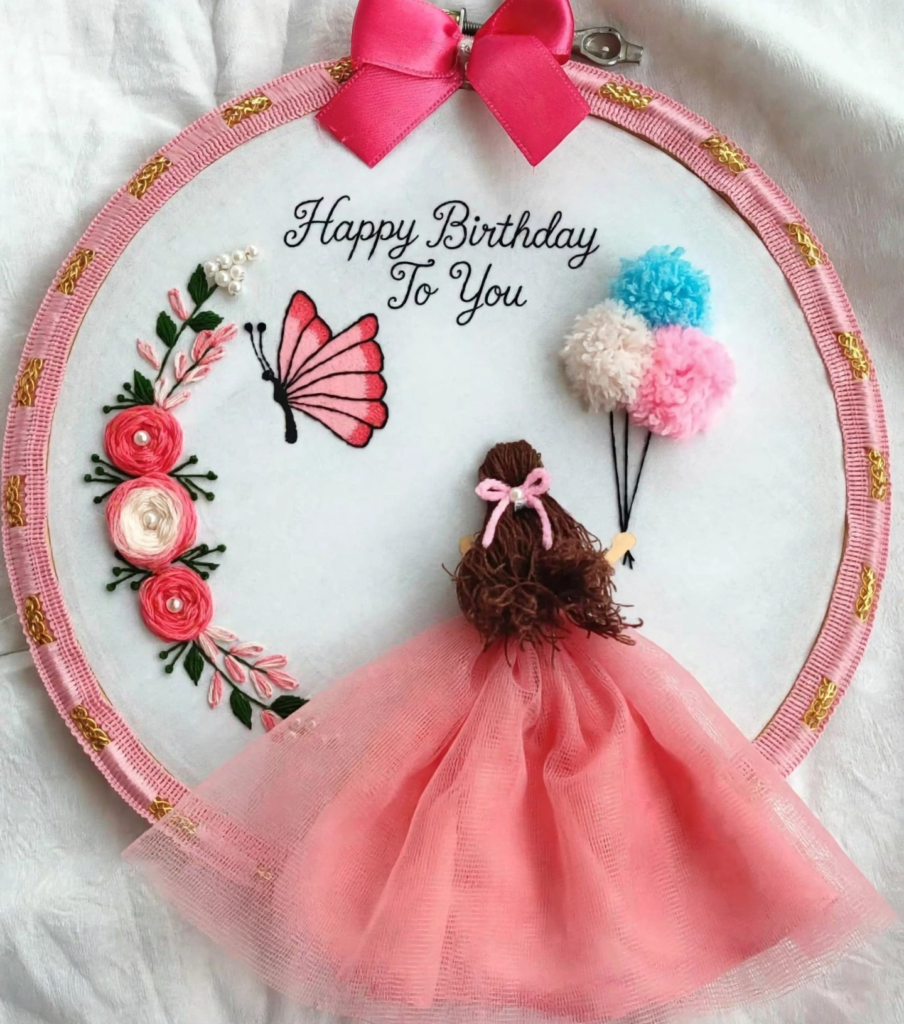 20 Retro Crafts to Try for a Creative Weekend Handmade embroidery hoop art featuring a 3D girl figure in a pink tulle dress and brown yarn hair holding pom-pom balloons, embroidered with "Happy Birthday To You," a pink butterfly, and floral accents, showcasing creative retro crafts.