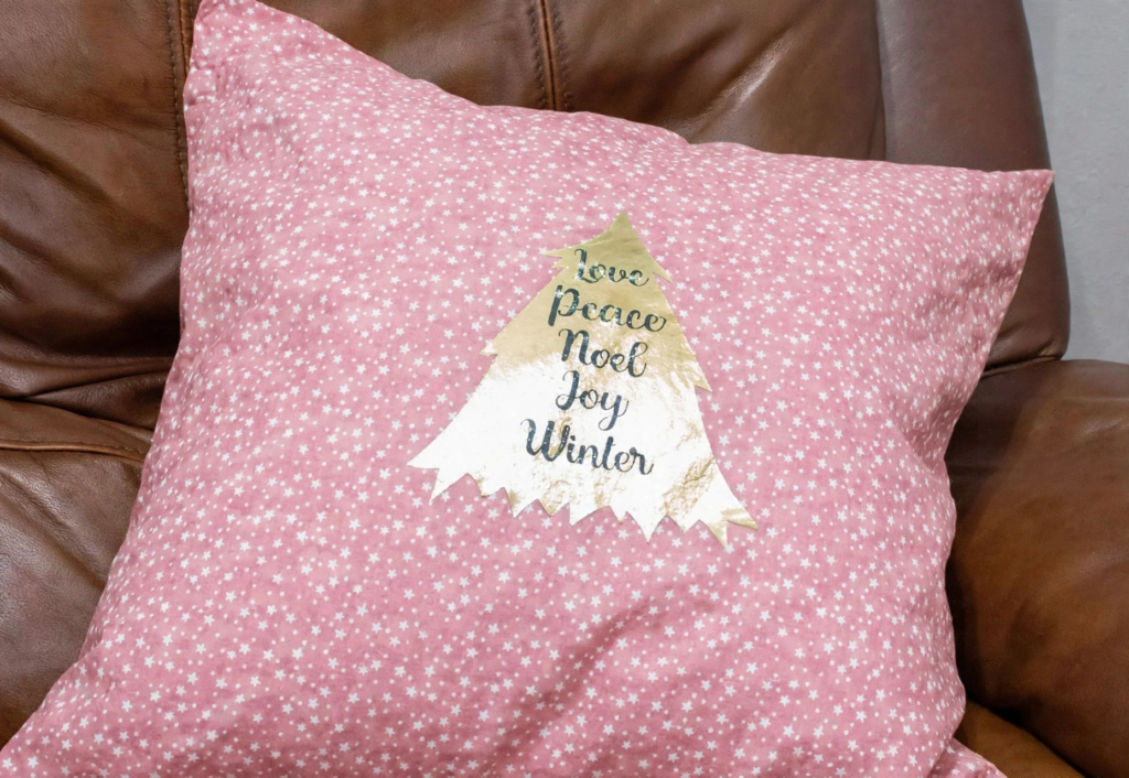 20 Retro Crafts to Try for a Creative Weekend Handmade pink floral print throw pillow resting on a brown leather armchair, decorated with a Christmas tree patch printed with holiday words, inspiring festive retro crafts home decor.
