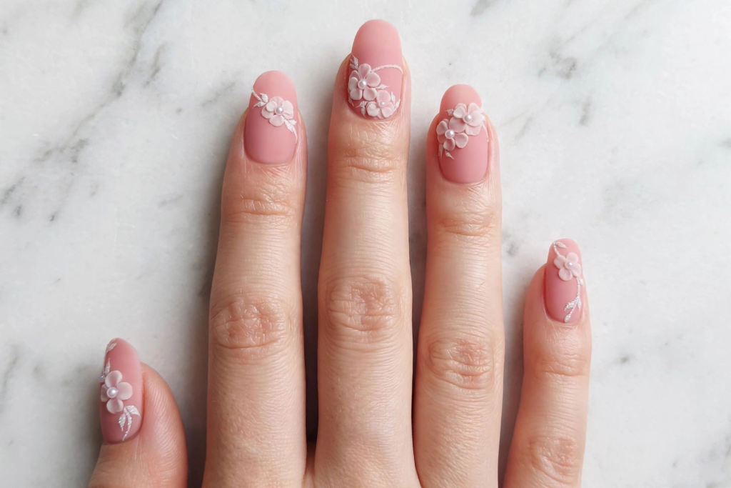 Hand showcasing easy 3D nail art designs featuring matte blush pink polish with delicate white sculpted acrylic flowers and pearl centers on a marble background.