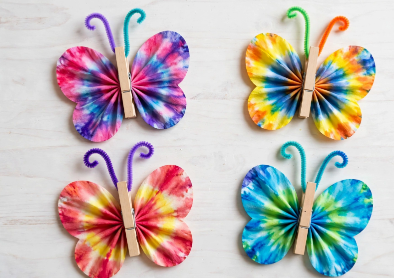 24 Preschool Seasonal Crafts for Spring, Summer, Fall & Winter Four colorful tie-dye coffee filter butterflies with wooden clothespin bodies and pipe cleaner antennae, an easy idea for preschool seasonal crafts.