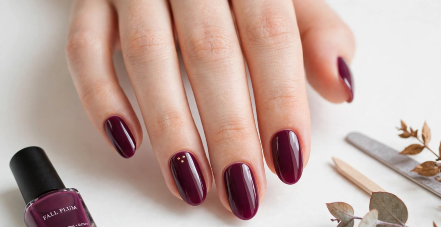 Glossy deep plum oval nails featuring delicate tiny gold dot accents on the ring finger alongside a matching polish bottle for fall plum nails 2026.