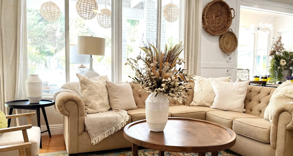 Sunlit living room showcasing cozy fall decor ideas for the home, featuring a tufted beige sectional sofa, woven wall baskets, neutral throw pillows, and a large dried wildflower and wheat arrangement in a ceramic vase on a round wooden coffee table.