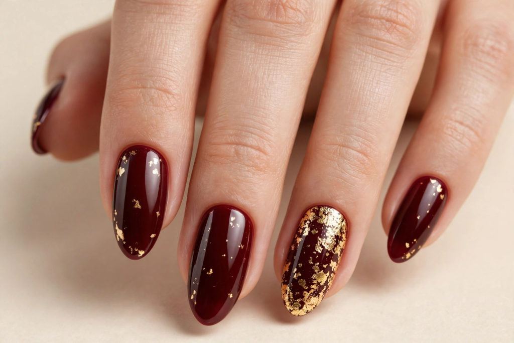 Fall nail inspo 2026 featuring glossy deep burgundy wine oval nails accented with shimmering gold foil flakes.   