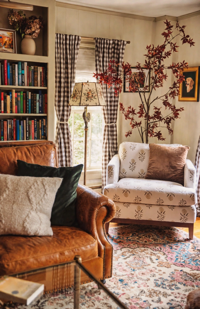 Cozy reading nook styled with fall home decor trends 2026, featuring a caramel leather armchair, gingham curtains, floral armchair, and an indoor autumn tree with red leaves.   