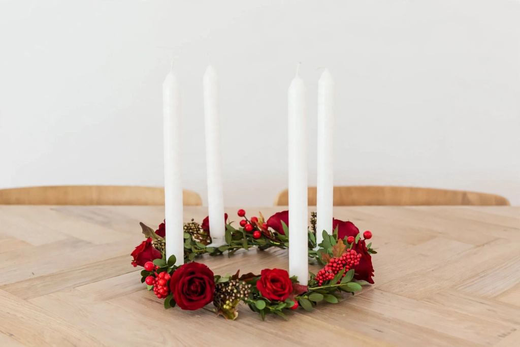 Four tall white taper candles in a floral wreath ring of red roses and berries on a wooden dining table for fall crafts for adults.